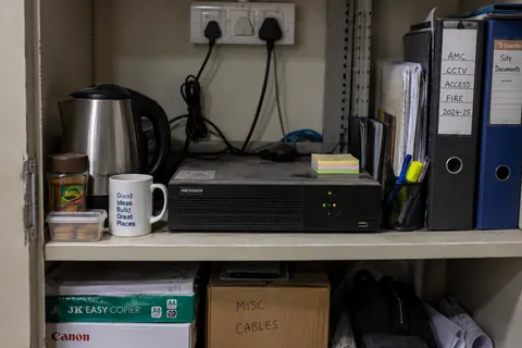 A digital recorder on a store cupboard shelf beside a kettle, a jar of coffee and a row of site files.