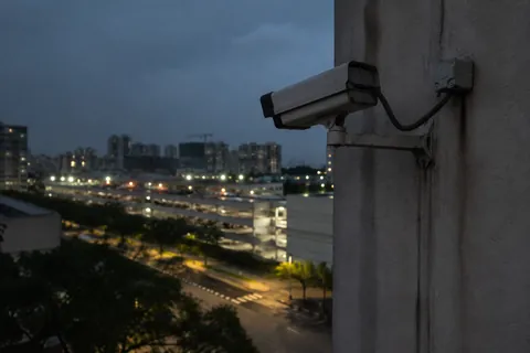 A camera on a column at dusk, looking out over a lit seafront road and the city behind it.