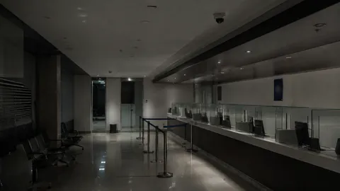 A closed bank branch after hours, the teller line unlit and the queue rail still in place along an empty customer floor.