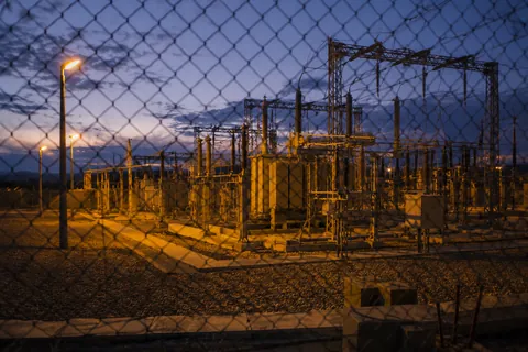 A substation at dusk seen through a chain-link fence, transformers and gantries lit from below.