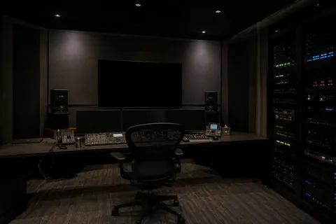 A broadcast control suite, the monitor wall dark and equipment racks lit along the back wall.