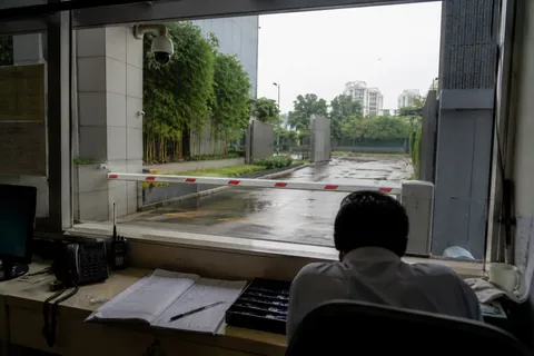 The view out from a gatehouse window: a guard at the desk with the visitor register and a handheld radio, the boom barrier down and the access road beyond it in the rain.