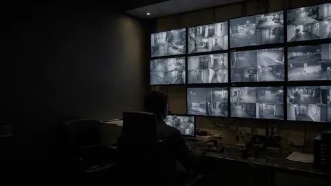 A darkened command room, a single operator facing a wall of camera monitors.