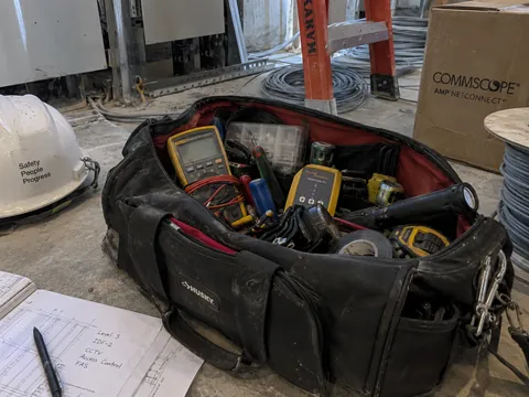 An open engineer's tool bag on a site floor, meters and hand tools inside it, a hard hat and a marked-up drawing beside it.